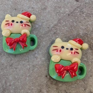 May include: Two green ceramic Christmas ornaments shaped like mugs with a cat wearing a Santa hat inside each mug. Each mug has a red bow.