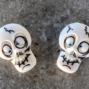 May include: Two white ceramic skull figurines with black painted details and blue eyes. The skulls have a stitched mouth and cracked forehead.