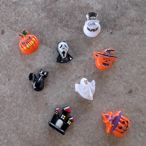 May include: A collection of Halloween-themed miniature figurines. Includes a black cat, a pumpkin, a ghost, a haunted house, a smiling snowman, and a Scream mask. The figurines are in various colors, including orange, black, and white.