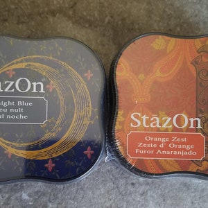 May include: Two ink pads, one blue and one orange, with the brand name "StazOn" printed on them. The blue ink pad has a crescent moon design and the text "Midnight Blue, Bleu nuit, Azul noche". The orange ink pad has a door knob design and the text "Orange Zest, Zeste d'Orange, Furor Anaranjado".