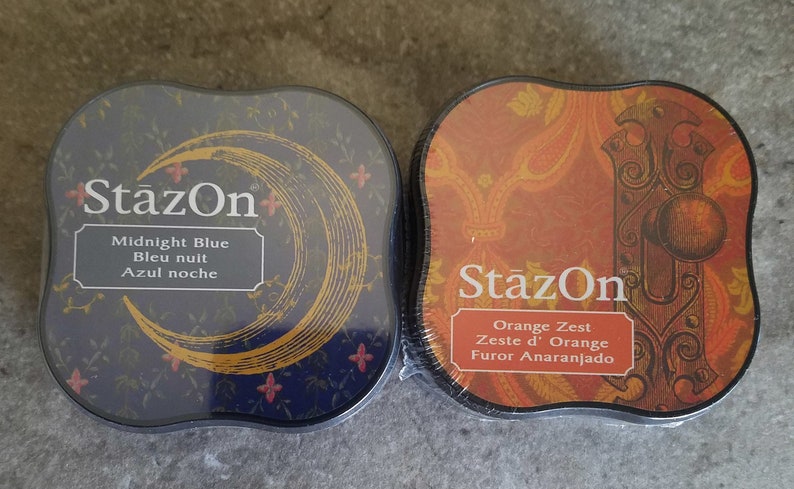 May include: Two ink pads, one blue and one orange, with the brand name "StazOn" printed on them. The blue ink pad has a crescent moon design and the text "Midnight Blue, Bleu nuit, Azul noche". The orange ink pad has a door knob design and the text "Orange Zest, Zeste d'Orange, Furor Anaranjado".
