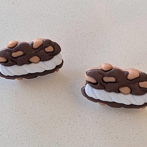May include: Two decorative charms shaped like ice cream sandwiches. The top and bottom layers are brown with tan-colored accents, and the middle layer is white. These charms are designed to be attached to shoes or other accessories.