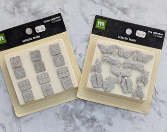 May include: Two packages of silver-colored metal stamp brads. The package on the left has 12 brads with designs including "LOVE" and a small house. The package on the right has 12 brads with designs including butterflies, dragonflies, and bugs.