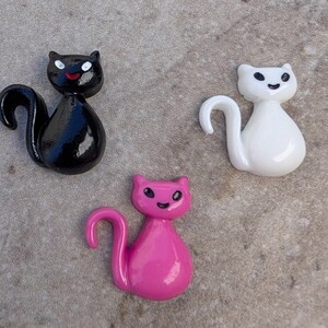 May include: Three ceramic cat figurines. One black cat, one white cat, and one pink cat. Each cat has a simple, cartoon-like face.