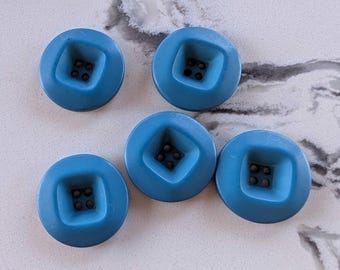 May include: Five round, light blue buttons with a square recessed center. Each button has four small holes in the center. The buttons are made of a smooth, solid material.