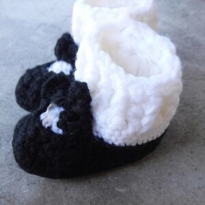 May include: A pair of handmade baby booties. The booties are black with white accents and a white cuff. The booties are made of a soft, textured yarn, perfect for a newborn.