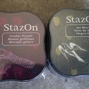 May include: Two ink pads, one black and one purple, with the brand name "StazOn" printed on the front. The black ink pad has a design of birds and leaves, and the purple ink pad has a peacock design. The ink pads are labeled with the ink color and name in English, French, and Spanish.