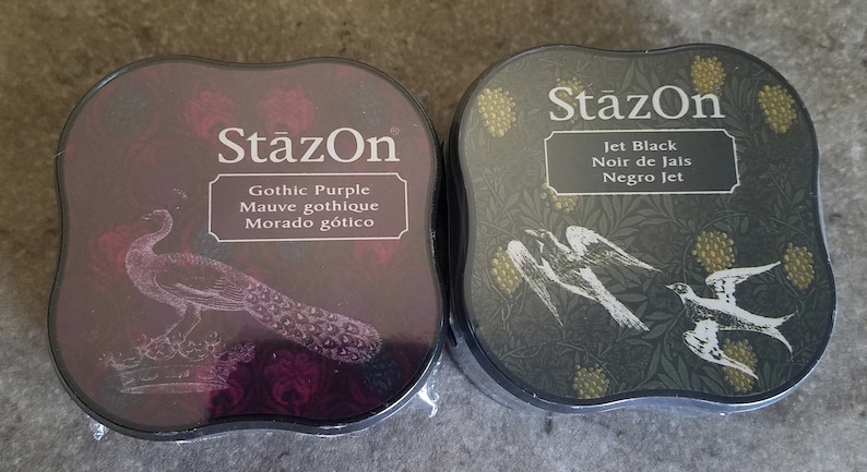 May include: Two ink pads, one black and one purple, with the brand name "StazOn" printed on the front. The black ink pad has a design of birds and leaves, and the purple ink pad has a peacock design. The ink pads are labeled with the ink color and name in English, French, and Spanish.