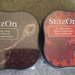 May include: Two round ink pads with black lids and a decorative design. The ink pad on the left has a brown background with a silver spoon and the text "StazOn Spiced Chai Chal &Eacute;pic&eacute; Chai Especiado". The ink pad on the right has a red background with a glass bottle and the text "StazOn St. Valentine Saint-Valentin San Valent&iacute;n".