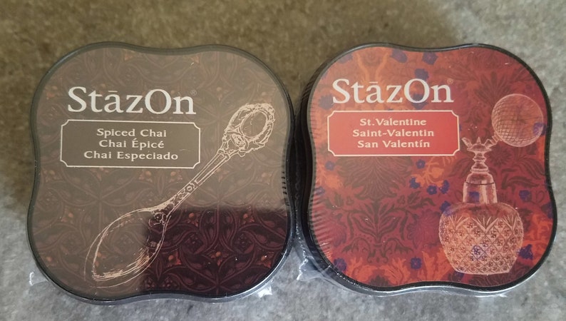 May include: Two round ink pads with black lids and a decorative design. The ink pad on the left has a brown background with a silver spoon and the text "StazOn Spiced Chai Chal &Eacute;pic&eacute; Chai Especiado". The ink pad on the right has a red background with a glass bottle and the text "StazOn St. Valentine Saint-Valentin San Valent&iacute;n".