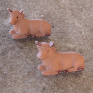 May include: Two brown ceramic figurines of cows, each with a brown and white face and horns. The cows are lying down with their legs tucked underneath them.