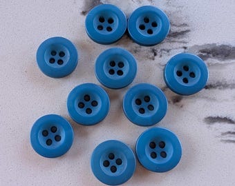 May include: A collection of ten round, teal-colored buttons with four holes each. The buttons are arranged on a light gray and white speckled surface. The buttons are made of a smooth, solid material.