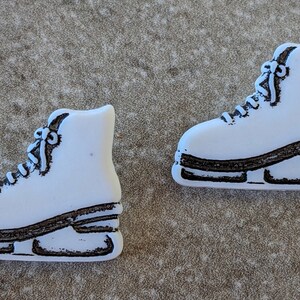 May include: Two white ice skate ornaments with black trim and laces are displayed on a textured, neutral-toned surface. The skates are side-by-side, showcasing their design. These decorative items are ideal for winter-themed crafts or holiday decor.