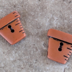 May include: Two terracotta-colored, decorative buttons in a flower pot shape. Each button has a keyhole-shaped buttonhole and black detailing. The buttons are angled and have a textured appearance, suitable for sewing or crafts.