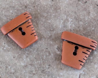 Puede incluir: Dos botones decorativos de color terracota con forma de maceta. Cada botón tiene un ojal en forma de cerradura y detalles en negro. Los botones están en ángulo y tienen un aspecto texturizado, adecuados para coser o manualidades.