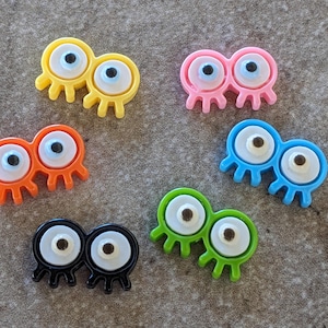 May include: Six colorful plastic toy monsters with googly eyes. Each monster has two eyes and four legs. The monsters are yellow, pink, orange, blue, green, and black.