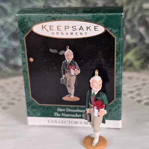 May include: A Hallmark Keepsake Ornament of Herr Drosselmeyer from The Nutcracker, featuring a figure in a dark green coat, maroon vest, and tan trousers, holding a gift. The ornament stands on a tan base in front of its original green box.