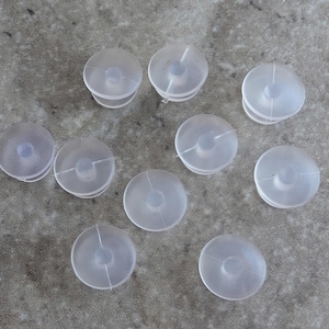 May include: A collection of nine translucent, circular plastic sewing machine bobbins. Each bobbin features a flat, disc-like shape with a central hub and a small, raised circular opening in the middle, designed for holding thread during sewing projects.