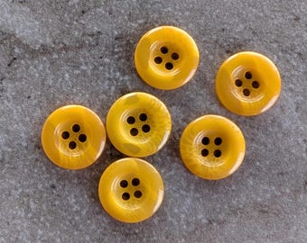 May include: Six round, yellow buttons with four holes each are arranged on a gray surface. The buttons have a marbled appearance, with variations in the yellow color. The buttons are a sewing or craft supply.