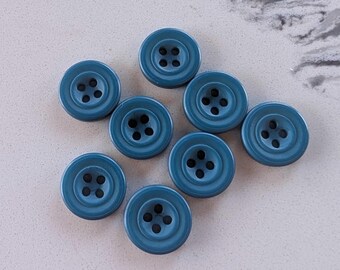 May include: A collection of eight teal-colored buttons. Each button is round with four holes in the center and a slightly raised outer rim. The buttons are arranged on a light gray surface.