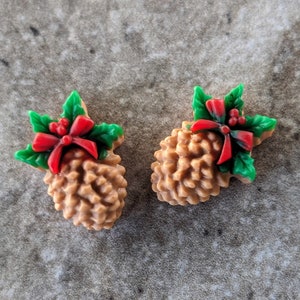 May include: Two brown pine cone ornaments with green holly leaves and red bows. The ornaments are perfect for Christmas decorations.