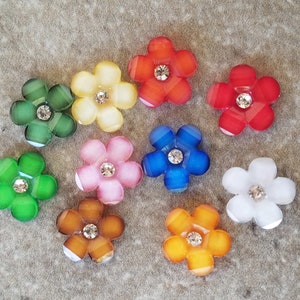 May include: Ten colorful flower-shaped buttons with a rhinestone center. The buttons are in shades of green, yellow, red, orange, pink, blue, and white.