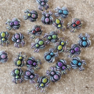 May include: A collection of colorful, glittery spider charms with various designs. The spiders are made of metal and have a 3D effect.