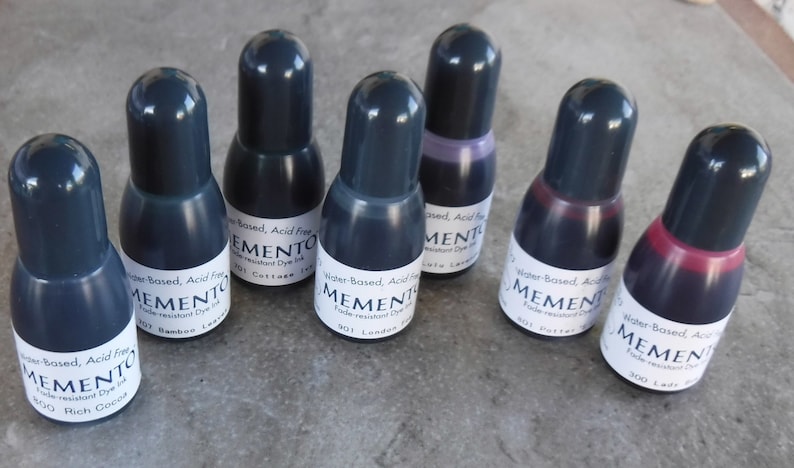 Puede incluir: Siete botellas de tinta Memento a base de agua, sin &aacute;cido y resistente a la decoloraci&oacute;n. Cada botella tiene una tapa gris oscuro y una etiqueta blanca con texto negro. Los colores de tinta var&iacute;an de gris oscuro a tonos de p&uacute;rpura y rojo.