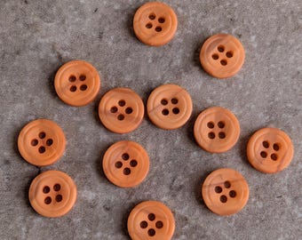 May include: A collection of eleven round, orange buttons, each featuring a smooth surface with a recessed center and four evenly spaced holes for attachment. The buttons have a consistent circular shape and a uniform matte orange color throughout.
