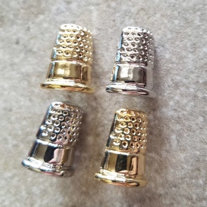May include: Four metal thimbles in gold and silver tones. The thimbles have a classic design with a rounded top covered in small dimples for gripping needles. The base of each thimble is smooth and shiny.