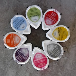 May include: A collection of ten Memento Dew Drop ink pads in various colors, including green, blue, red, orange, yellow, and gray. Each pad features the Memento Dew Drop logo and the ink color name in English and another language.