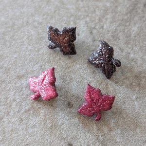 May include: Four stud earrings shaped like ivy leaves. Two earrings are a dark brown color with a glittery finish, and two earrings are a vibrant pink color with a glittery finish. Each leaf-shaped earring features a distinct three-lobed design.
