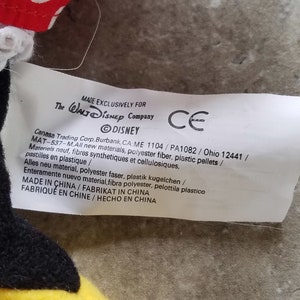 Może przedstawiać: Biała etykieta z czarnym tekstem, kt&oacute;ry brzmi "MADE EXCLUSIVELY FOR The WALT Disney Company" i "DISNEY" w k&oacute;łku. Etykieta zawiera r&oacute;wnież tekst "Canasa Trading Corp, Burbank, CA:ME 1104/PA1082 / Ohio 12441/ MAT-537-M.All new materials, polyester fiber, plastic pellets / Materiels neuf, fibres synthetiques et cellulosiques, pastilles en plastique / Alles neu material, polyester faser, plastik kugelchen / Enteramente nuevo material, fibra polyester, pelottila plastico MADE IN CHINA/FABRIKAT IN CHINA FABRIQU&Eacute; EN CHINE/HECHO EN CHINA" oraz znak CE.