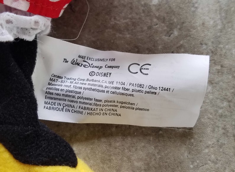Może przedstawiać: Biała etykieta z czarnym tekstem, kt&oacute;ry brzmi "MADE EXCLUSIVELY FOR The WALT Disney Company" i "DISNEY" w k&oacute;łku. Etykieta zawiera r&oacute;wnież tekst "Canasa Trading Corp, Burbank, CA:ME 1104/PA1082 / Ohio 12441/ MAT-537-M.All new materials, polyester fiber, plastic pellets / Materiels neuf, fibres synthetiques et cellulosiques, pastilles en plastique / Alles neu material, polyester faser, plastik kugelchen / Enteramente nuevo material, fibra polyester, pelottila plastico MADE IN CHINA/FABRIKAT IN CHINA FABRIQU&Eacute; EN CHINE/HECHO EN CHINA" oraz znak CE.