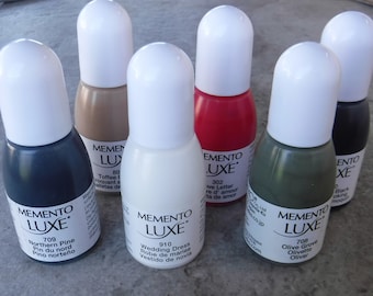 May include: A collection of ten ink bottles in various colors, including black, red, white, green, and brown. Each bottle has a white cap and a label with the brand name "Memento Luxe" and the color name in English and French.
