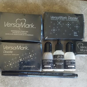 May include: A set of VersaMark watermark stamp pads and ink refills in black packaging. The pads include "Dazzle" in champagne and "Frost" colors. The ink refills are also in champagne and frost colors. The set also includes a black VersaMarker pen.