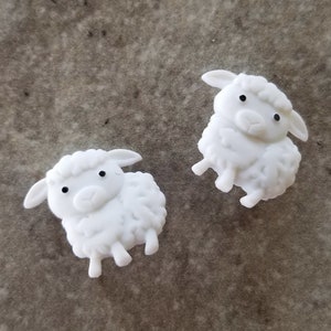 May include: Two white sheep figurines with black eyes and detailed wool textures. The sheep are in a playful pose, with their legs slightly bent. The background is a neutral, textured surface.