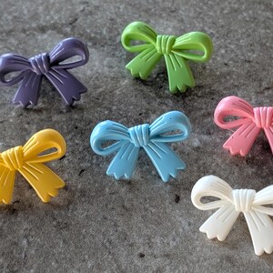 May include: A collection of six bow-shaped buttons in various solid colors including purple, light green, yellow, light blue, pink, and white. Each button features a molded ribbon design with a central knot and two dangling ends with notched edges.