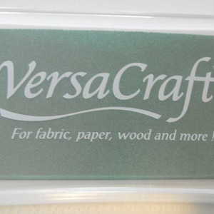 May include: A clear plastic container with a green ink pad inside. The ink pad has the text "VersaCraft" in white lettering and the words "For fabric, paper, wood and more!" in white lettering.