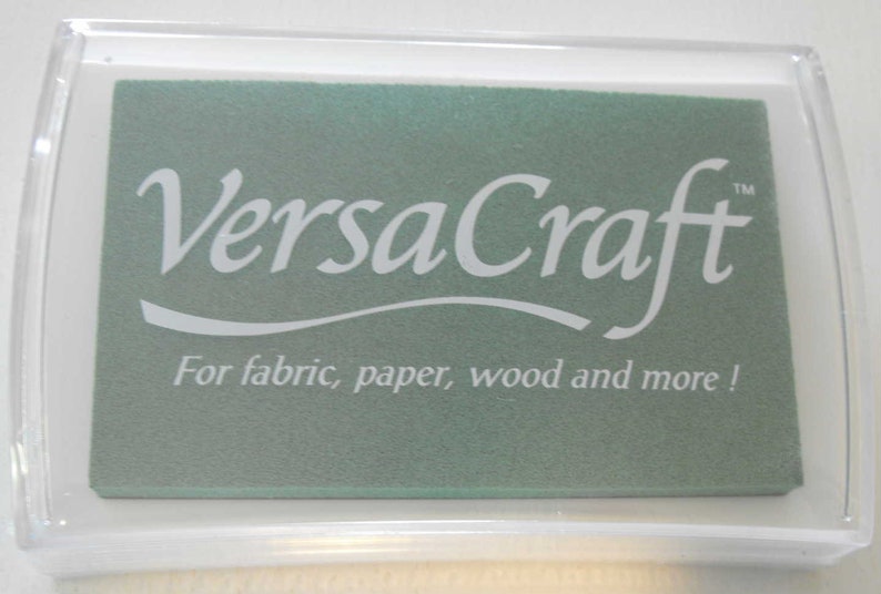 May include: A clear plastic container with a green ink pad inside. The ink pad has the text "VersaCraft" in white lettering and the words "For fabric, paper, wood and more!" in white lettering.