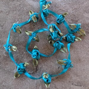 May include: A necklace made of teal and gold ribbon roses. The teal ribbon forms the stems and the roses are made of teal and gold ribbon. The necklace is lying on a brown surface.