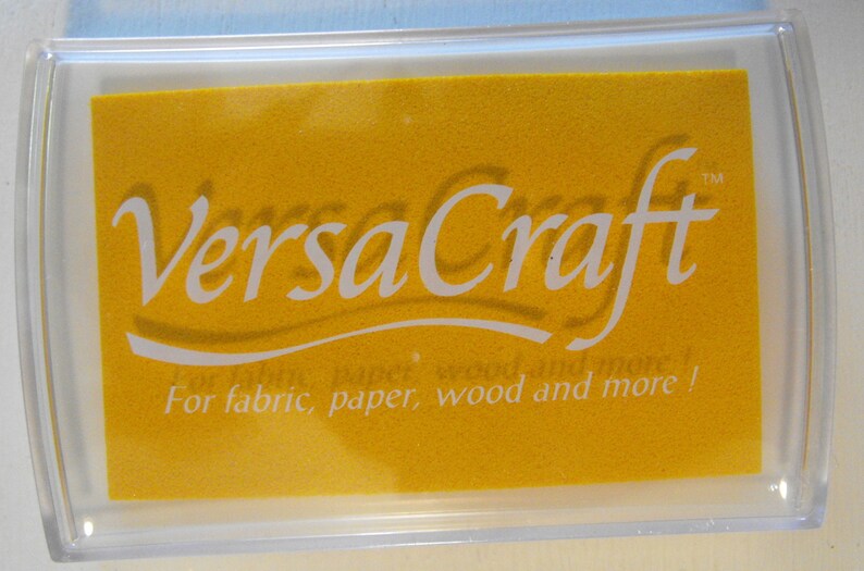 May include: A yellow ink pad with the text "VersaCraft" in white lettering. The ink pad is in a clear plastic case and has the text "For fabric, paper, wood and more!" in white lettering.