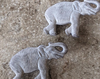 May include: Two gray elephant figurines with detailed carved textures. The elephants are in a walking pose, with trunks raised. The background is a mottled, speckled surface.