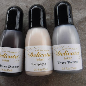 Op de afbeelding: Drie flesjes Delicata Inkt in de kleuren Donkerbruin Schimmer, Champagne en Zilveren Schimmer. Elk flesje bevat 0,5 fl oz (15 ml).