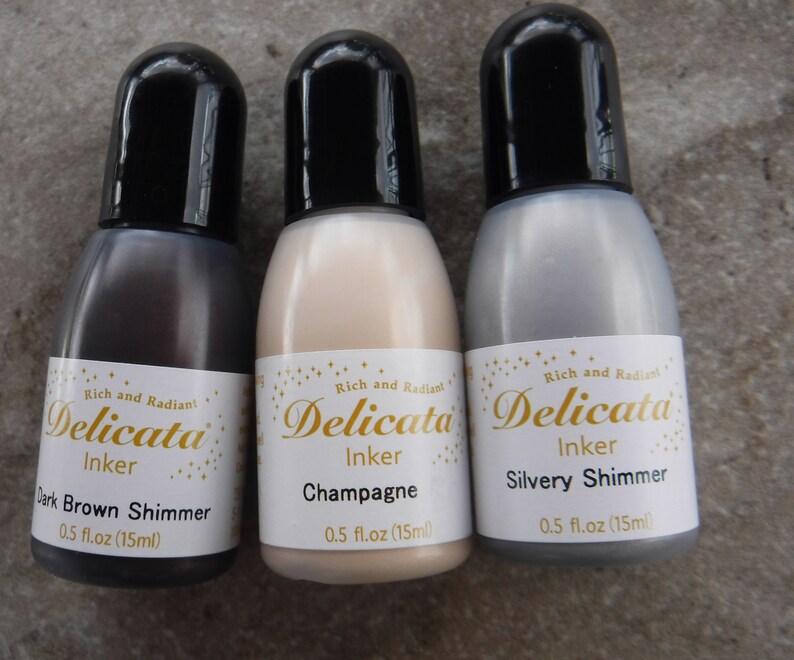 Op de afbeelding: Drie flesjes Delicata Inkt in de kleuren Donkerbruin Schimmer, Champagne en Zilveren Schimmer. Elk flesje bevat 0,5 fl oz (15 ml).
