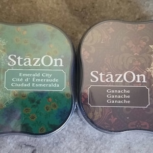 May include: Two ink pads with decorative designs. The left pad is green with a gold lantern design and the text "StazOn Emerald City Cit&eacute; d'&Eacute;meraude Ciudad Esmeralda". The right pad is brown with a gold clock design and the text "StazOn Ganche Ganche Ganche".