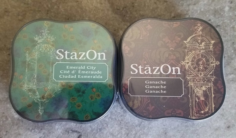 May include: Two ink pads with decorative designs. The left pad is green with a gold lantern design and the text "StazOn Emerald City Cit&eacute; d'&Eacute;meraude Ciudad Esmeralda". The right pad is brown with a gold clock design and the text "StazOn Ganche Ganche Ganche".