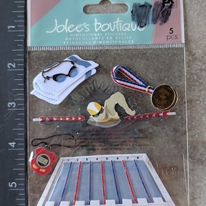 May include: A package of dimensional stickers featuring swimming-themed designs. Includes a swimmer, a stopwatch, a medal, a pool, goggles, and a towel. The package reads "Jolee's Boutique" and "5 pcs".