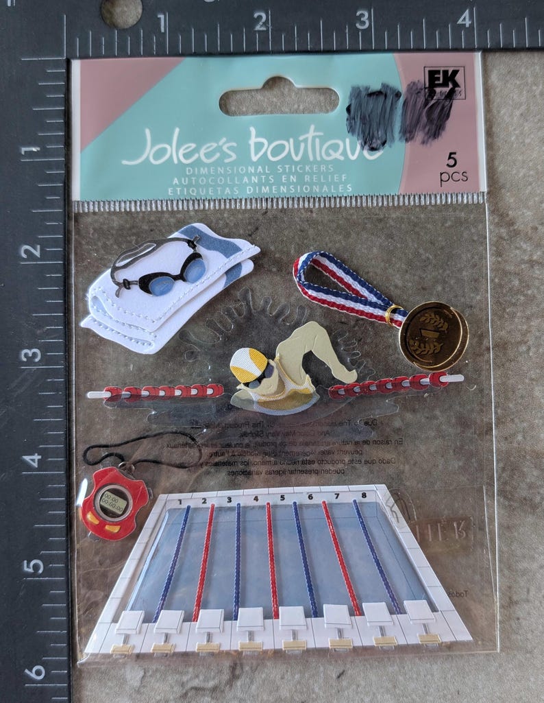 May include: A package of dimensional stickers featuring swimming-themed designs. Includes a swimmer, a stopwatch, a medal, a pool, goggles, and a towel. The package reads "Jolee's Boutique" and "5 pcs".