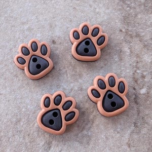 May include: Four individual buttons shaped like animal paw prints. Each button features a light tan outer border and a dark brown center pad with four smaller dark brown toe pads. There are two circular holes in the center of each paw print button.
